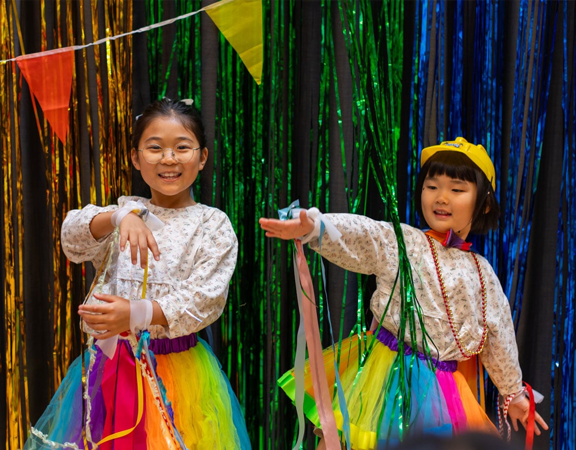Two children in bright dresses joyfully dancing in front of a colorful backdrop.