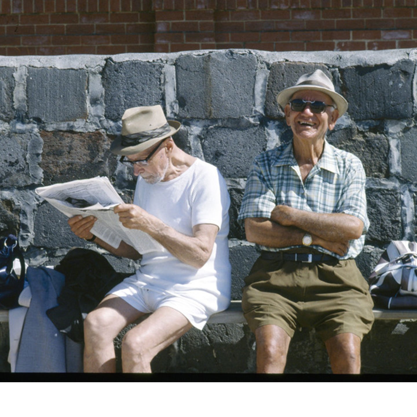 Two old men sitting on a bench, one is reading a newspaper and the other is smiling at the camera