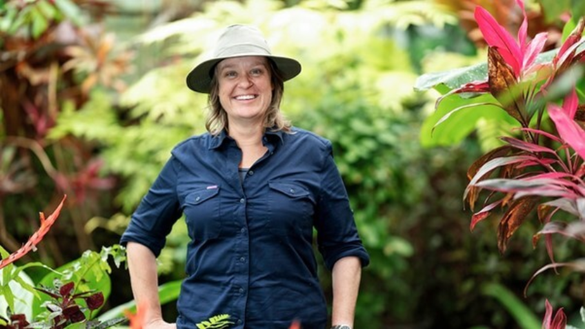 Woman wearing button up navy shirt and hat, smiling at camera. Behind her are a lot of plants.