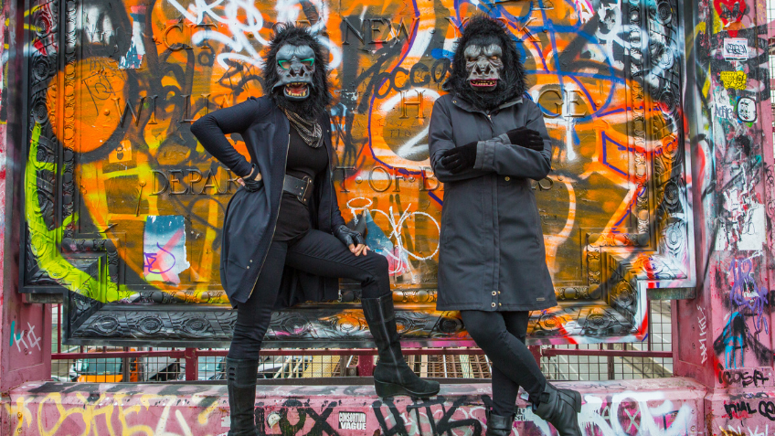 Two women in black trench coats leaning on a graffiti-ed wall 