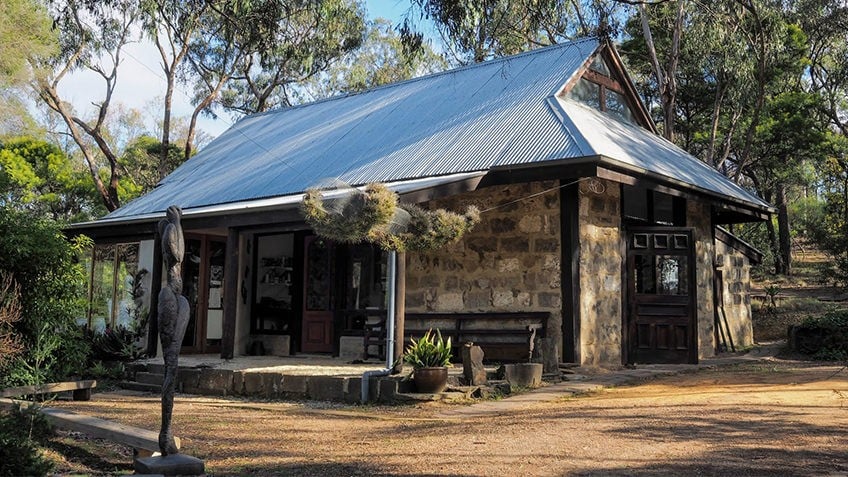 stone bush cottage with corrugated-iron roof surrounded by gum trees and statues