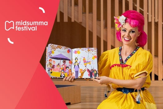 Woman with hot pink hair and yellow dress smiling at the camera, with a storybook opened and facing the camera.