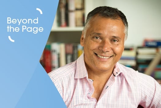 Man in baby pink button up shirt smiling at camera, with books in background