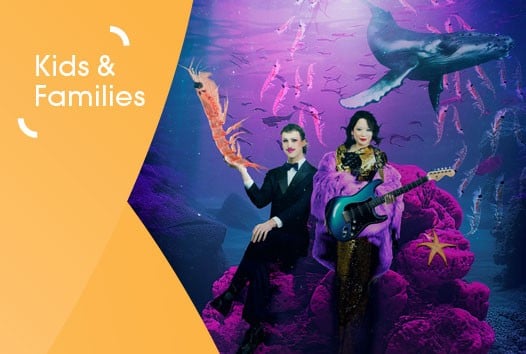 Woman in sparkly dress holding guitar and man in tuxedo holding krill underwater. Whale in the background